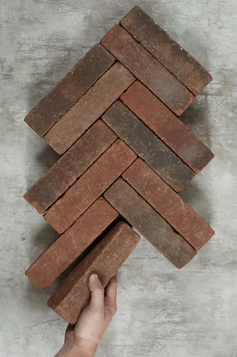Hand adds 12th brick to 11 Antique Red Clay Pavers arranged on grey background in herringbone pattern. Free UK delivery available.