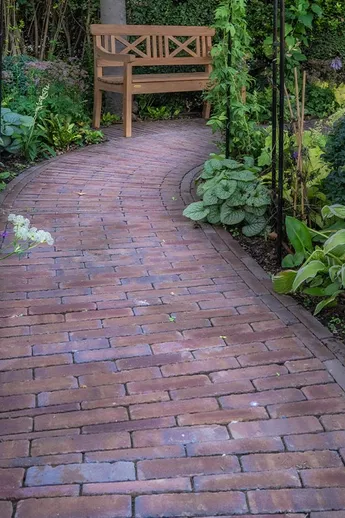 Path of Abbey Dark Multi Brick Pavers curves between beds of hostas and shrubs, past a wooden bench. Design by Jo Thompson.
