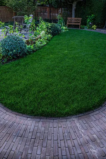 Sinuous path of Abbey Dark Multi clay pavers encircles lawn and bed, rising to patio with wooden seats. Build by Shoots and Leaves.