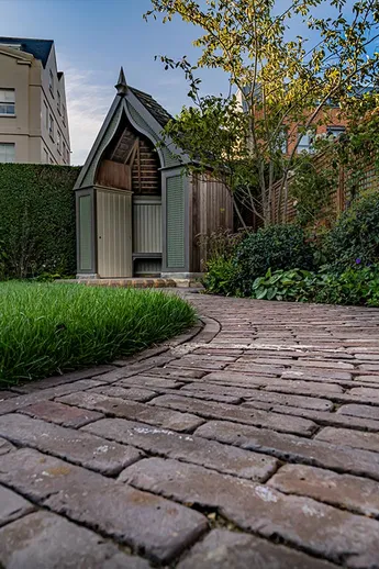 Curved path of Abbey Dark Multi brick pavers laid between lawn and bed leads to ogee-arched summer house and gate in hedge.