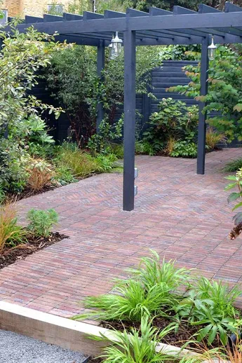 2 steps up to area paved with Bexhill Brick Pavers, covered by dark grey wooden pergola, surrounded by angular planted borders.