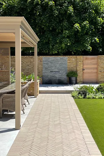 Stone Grey Brick Pavers laid between artificial lawn and dining area under wooden pergola against house. Design by Tom Howard.